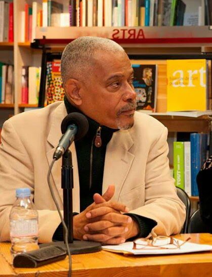 Robert Berrouët-Oriol à une table de rencontre, devant des rayonnages de livres
